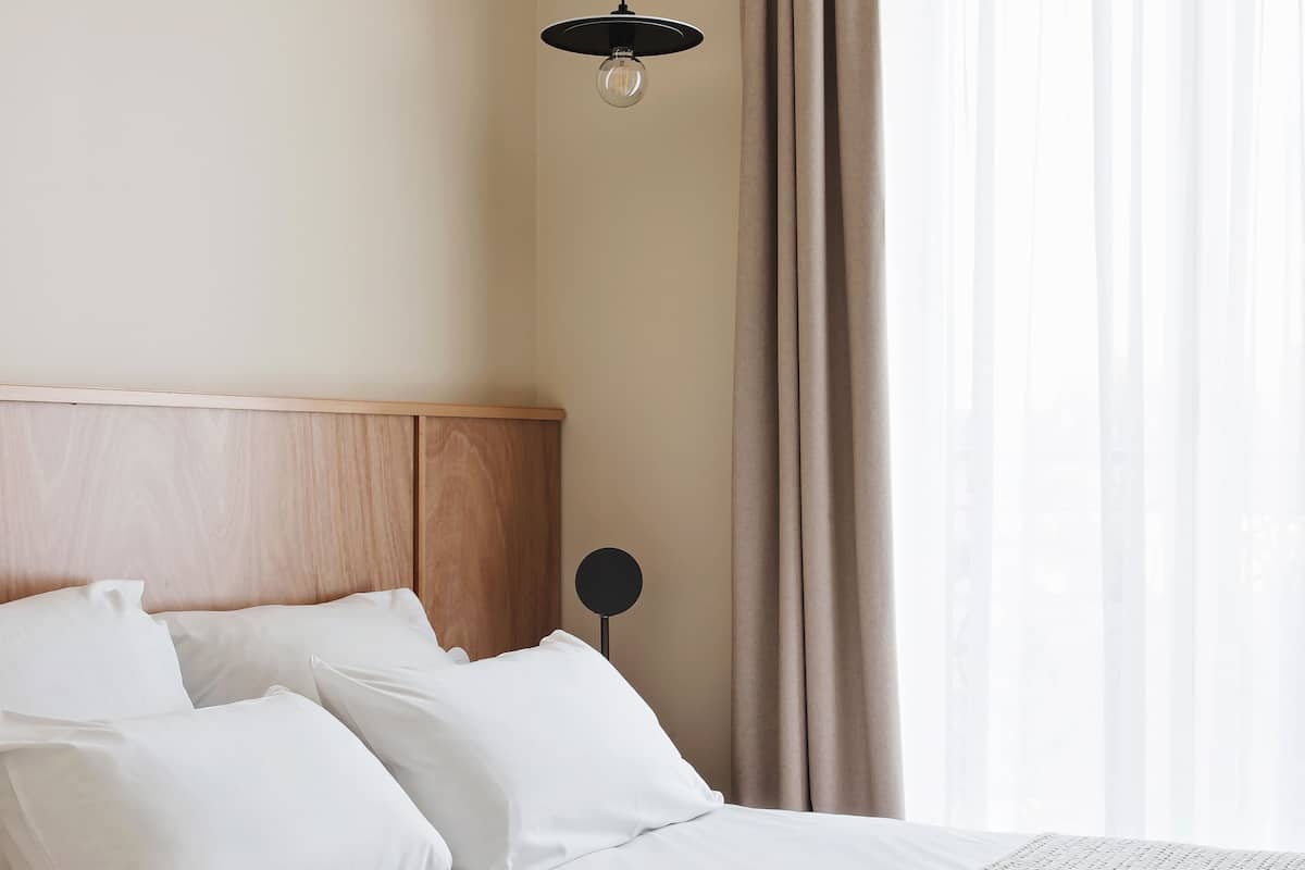 A hotel room with a bed, white bedding, a wooden headboard, a hanging light fixture, and sheer curtains.
