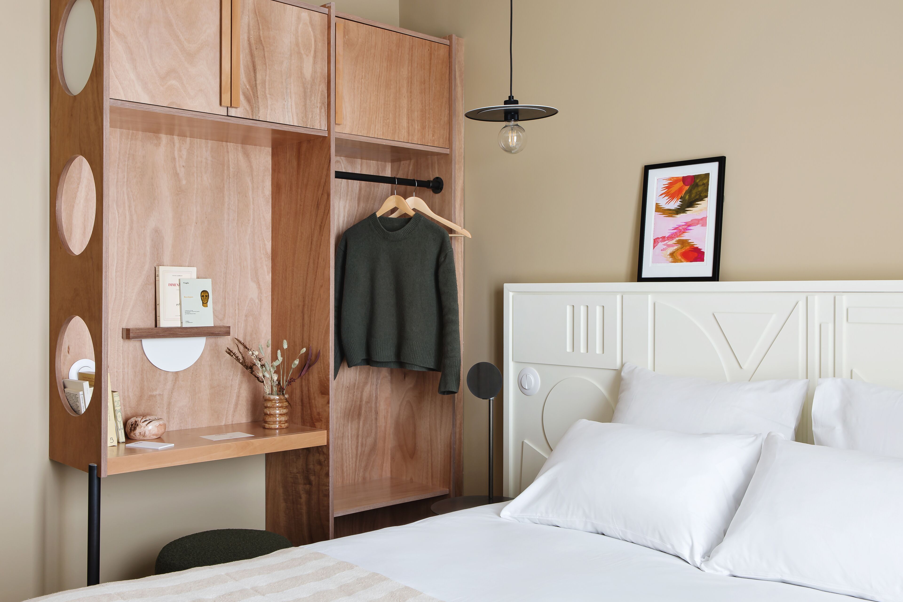 A bedroom with a bed, white pillows, a hanging robe, a wooden wardrobe, a small shelf with books and a vase, and a framed picture on the wall.