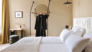 A neatly arranged bedroom with a bed, pillows, a hanging coat rack, a bedside table with a vase, and a circular mirror on the wall. - Hôtel Orphée - Orso hotel (Paris)