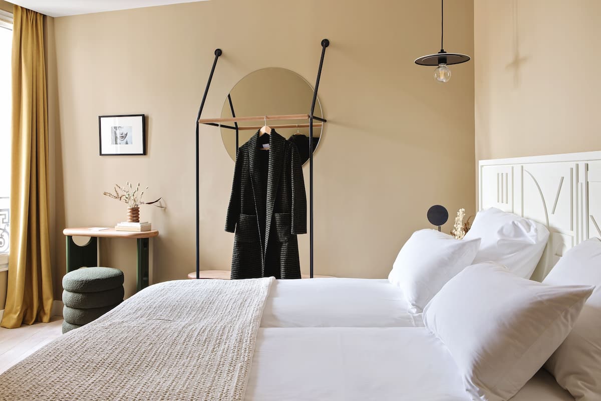A neatly arranged bedroom with a bed, pillows, a hanging coat rack, a bedside table with a vase, and a circular mirror on the wall.
