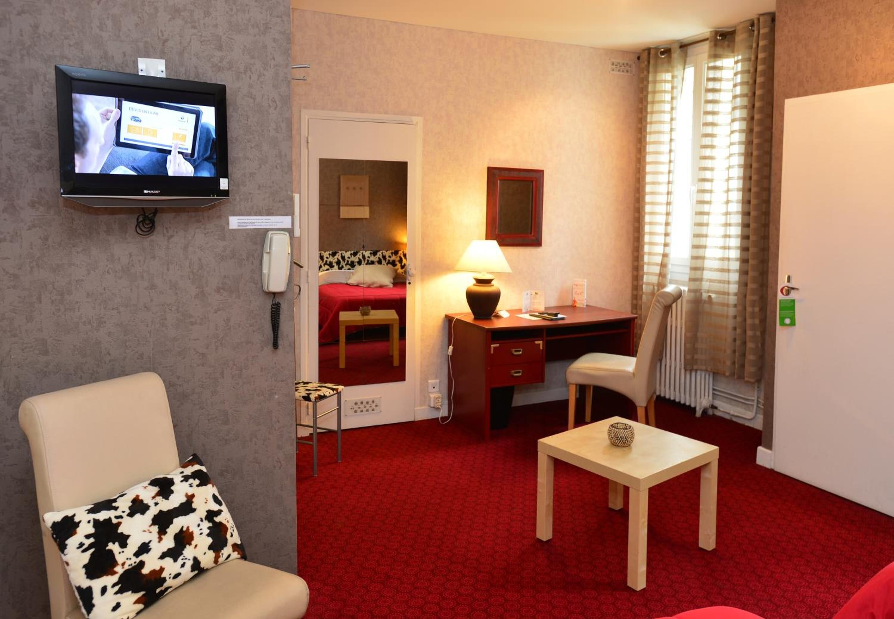 Una habitación de hotel con alfombra roja, un escritorio con lámpara, una silla, un televisor montado en la pared y una cama con cubierta roja.