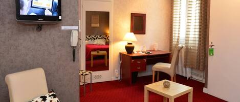 A hotel room with a red carpet, a desk with a lamp, a chair, a television mounted on the wall, and a bed with a red cover.