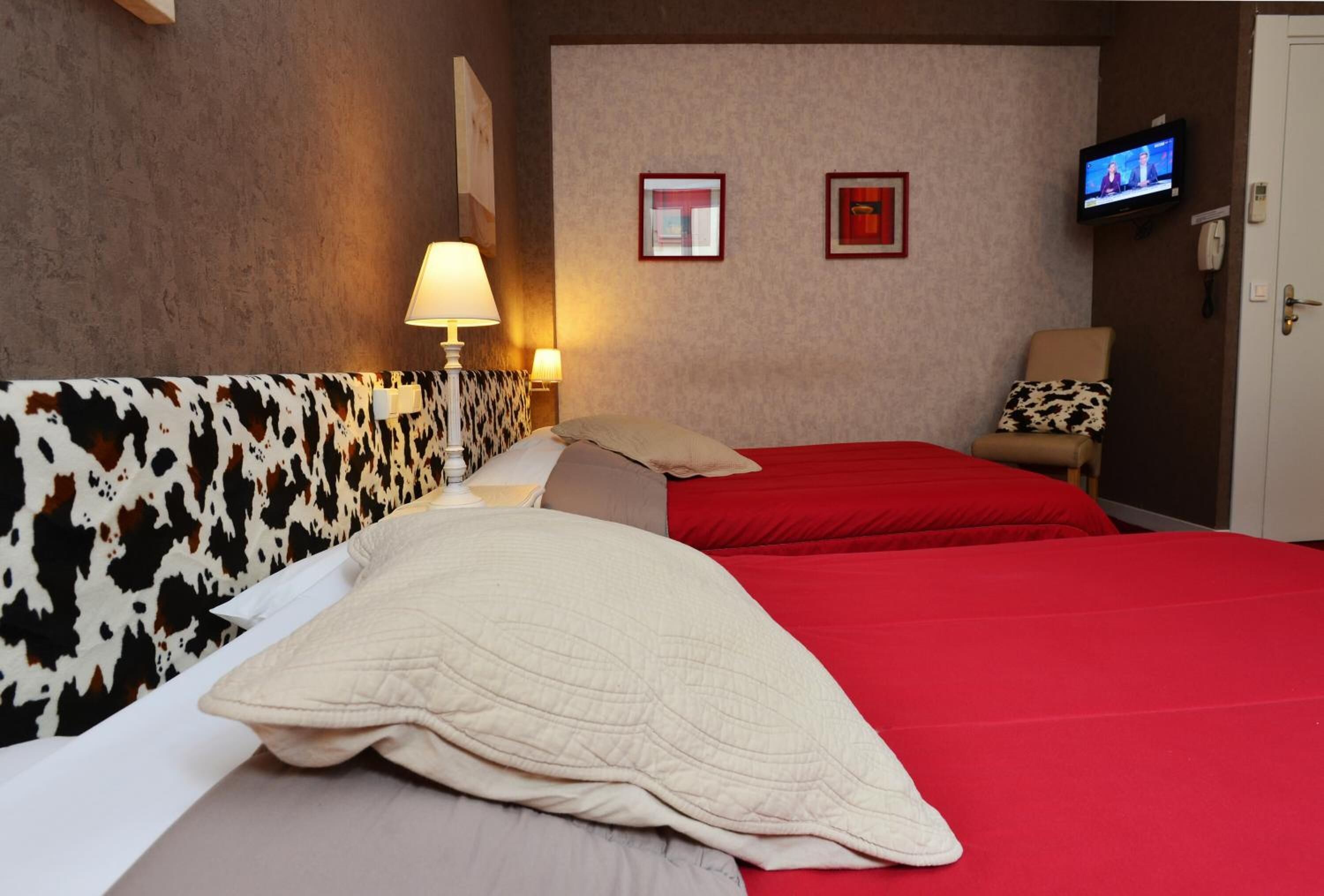 A hotel room with a cow print headboard, two red beds, a television on the wall, and a chair with a patterned cushion.
