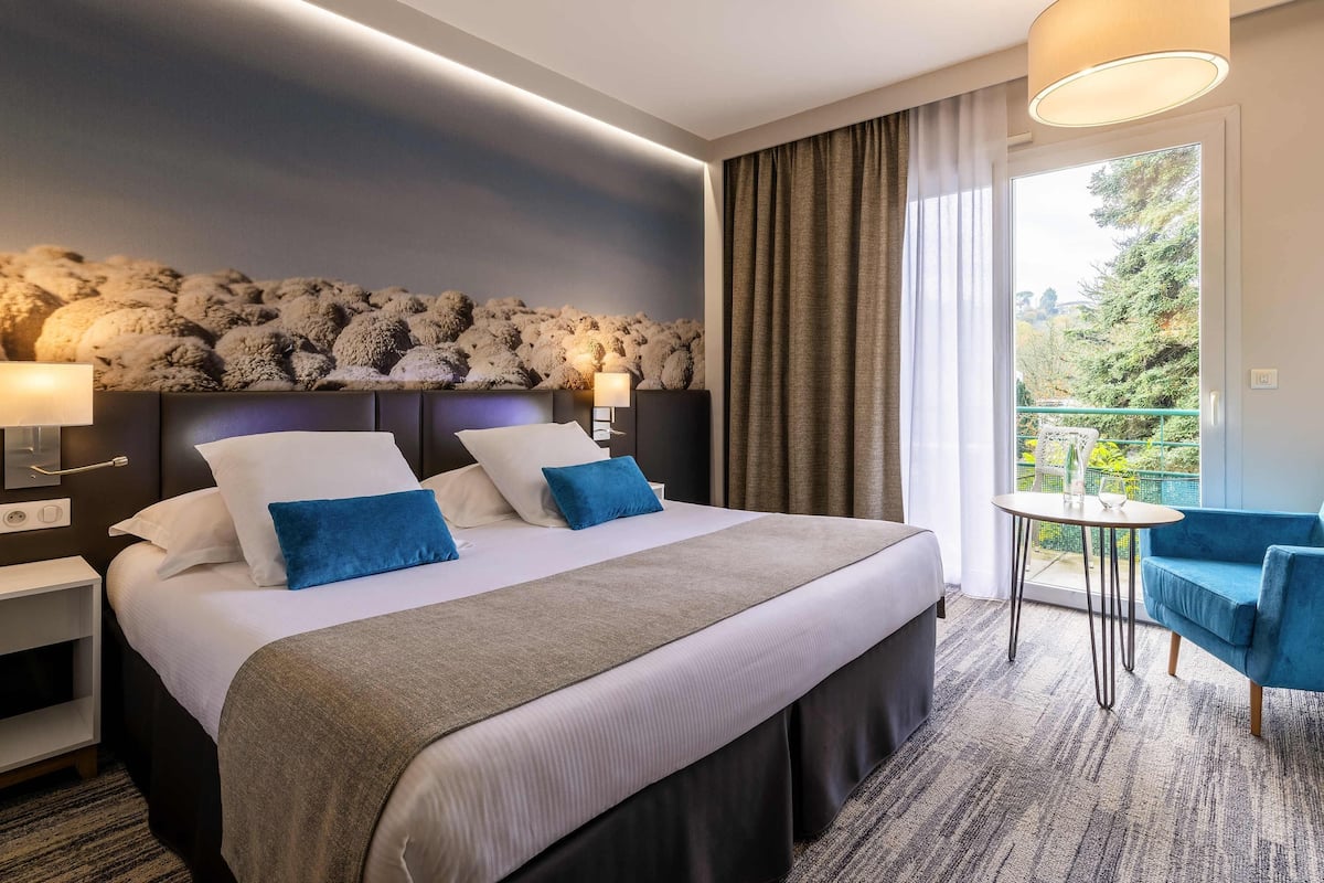 A hotel room with a bed, bedside lamps, a view of greenery, a small balcony with a table and chairs, and a decorative wall.