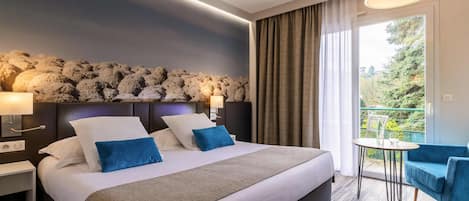 Habitación de hotel con cama, lámparas de noche, vistas a zonas verdes, un pequeño balcón con mesa y sillas, y un detalle decorativo en la pared.