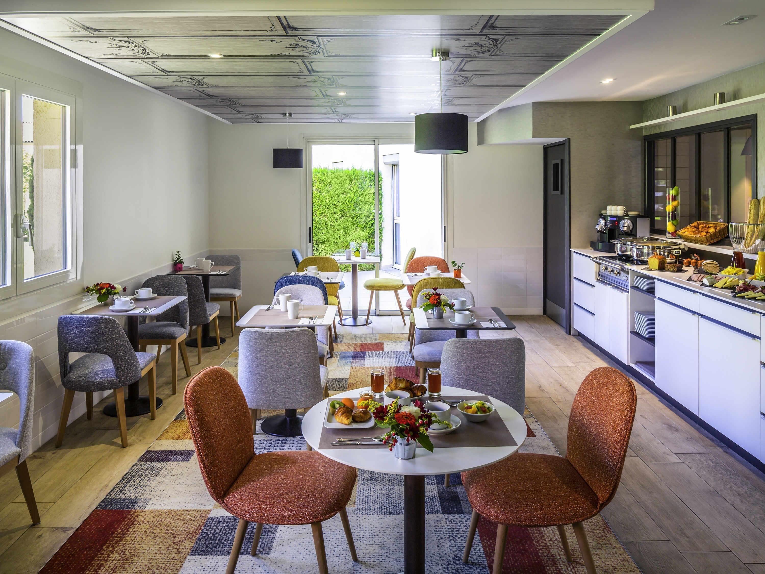 A modern dining area with a central round table, surrounded by grey upholstered chairs. A kitchen area with a buffet setup is visible to the right.