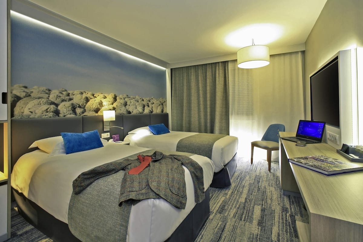 A hotel room with two beds, a desk with a laptop, a TV, and a large window with curtains.