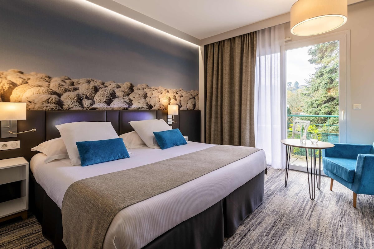 A hotel room with a bed, bedside lamps, a view of greenery, and a balcony with chairs.