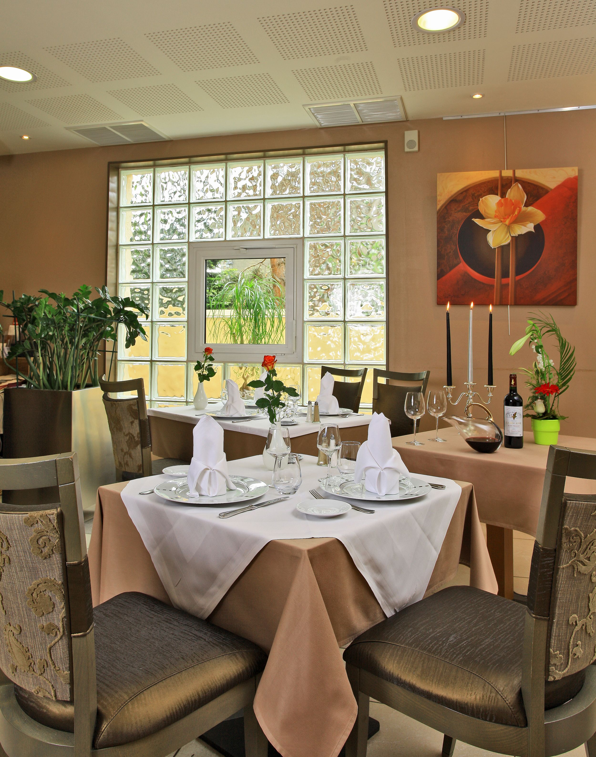 A dining area with a table set for four, featuring white tablecloths, plates, glasses, and cutlery. There are potted plants and a painting on the wall.