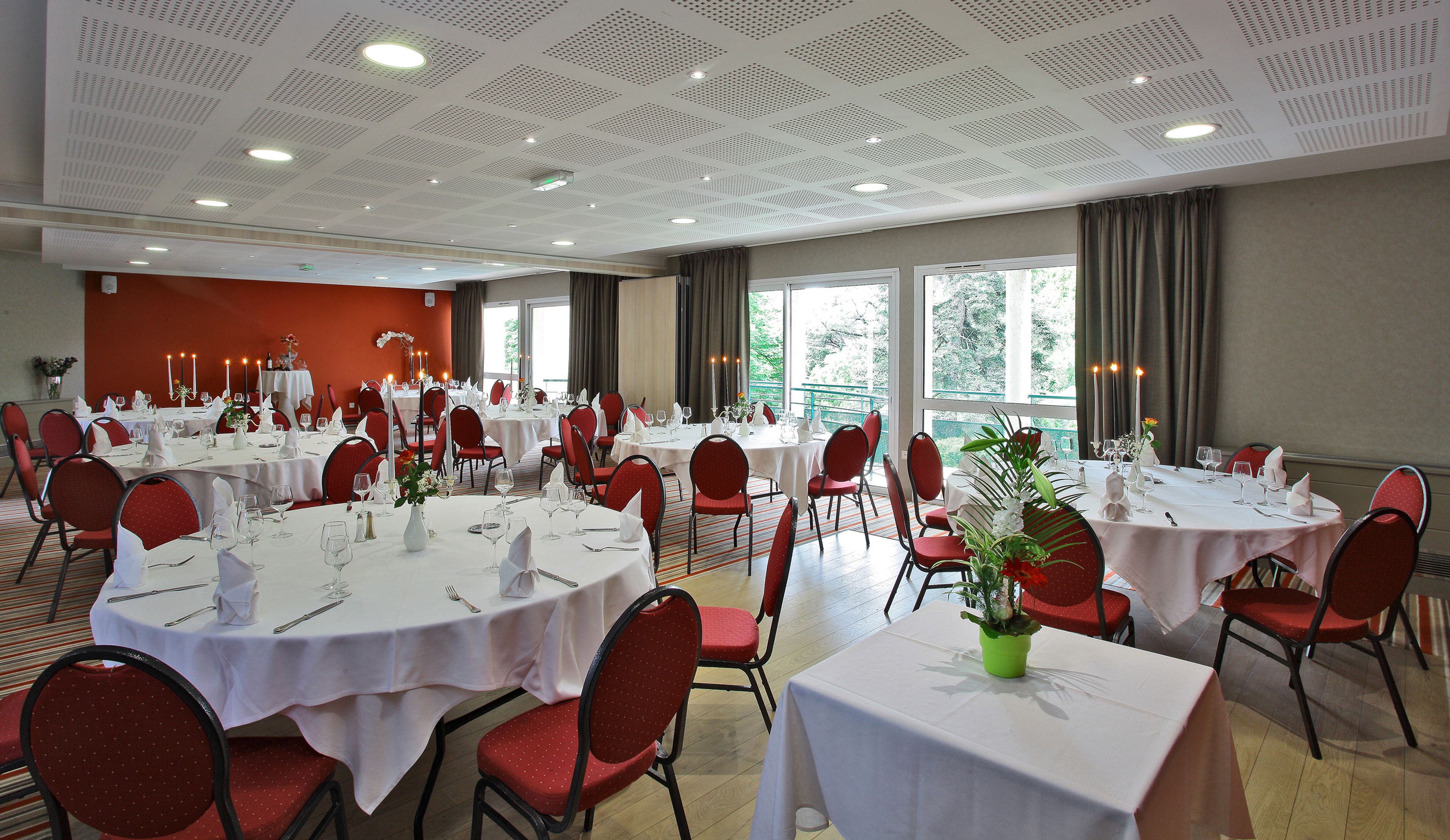 A banquet hall with round tables set for dining, red chairs, and large windows offering a view of greenery.