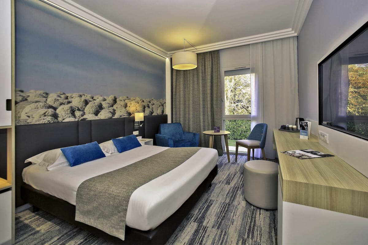 A hotel room with a large bed, a desk, a chair, a small table, and a window with a view of trees.