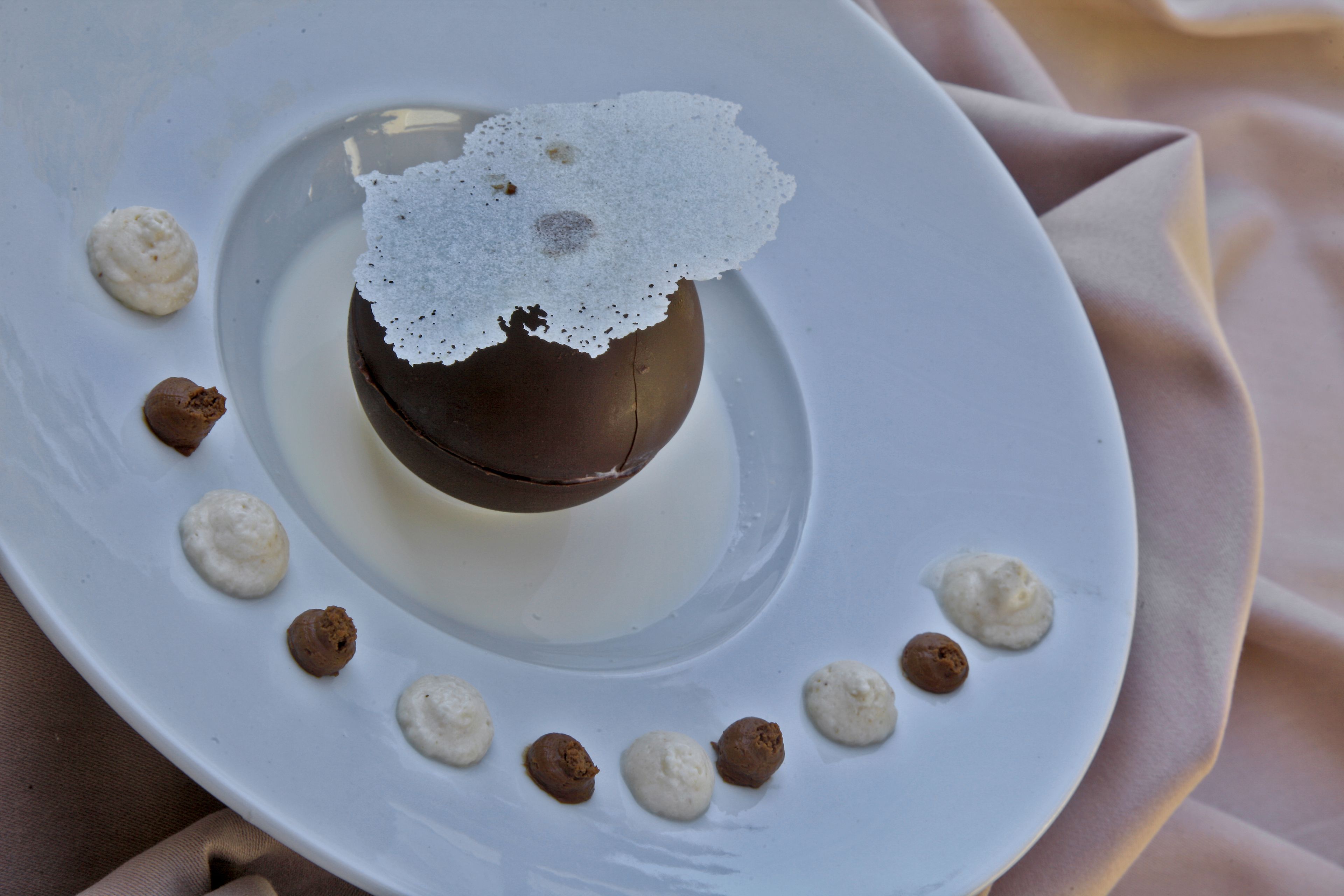A dessert on a white plate with a chocolate sphere and white cream dots around it.