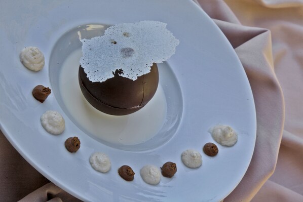 A dessert on a white plate with a chocolate sphere and white cream dots around it.
