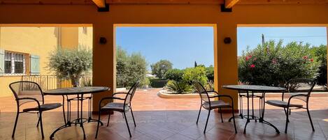 A covered patio with four tables and chairs, a view of the garden, and a yellow building with a window.