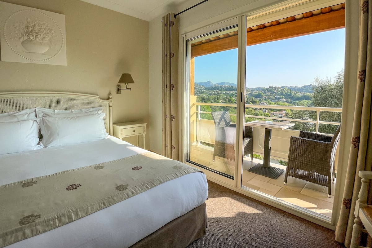 A hotel room with a bed, a nightstand, a wall-mounted lamp, and a sliding glass door leading to a balcony with outdoor furniture.