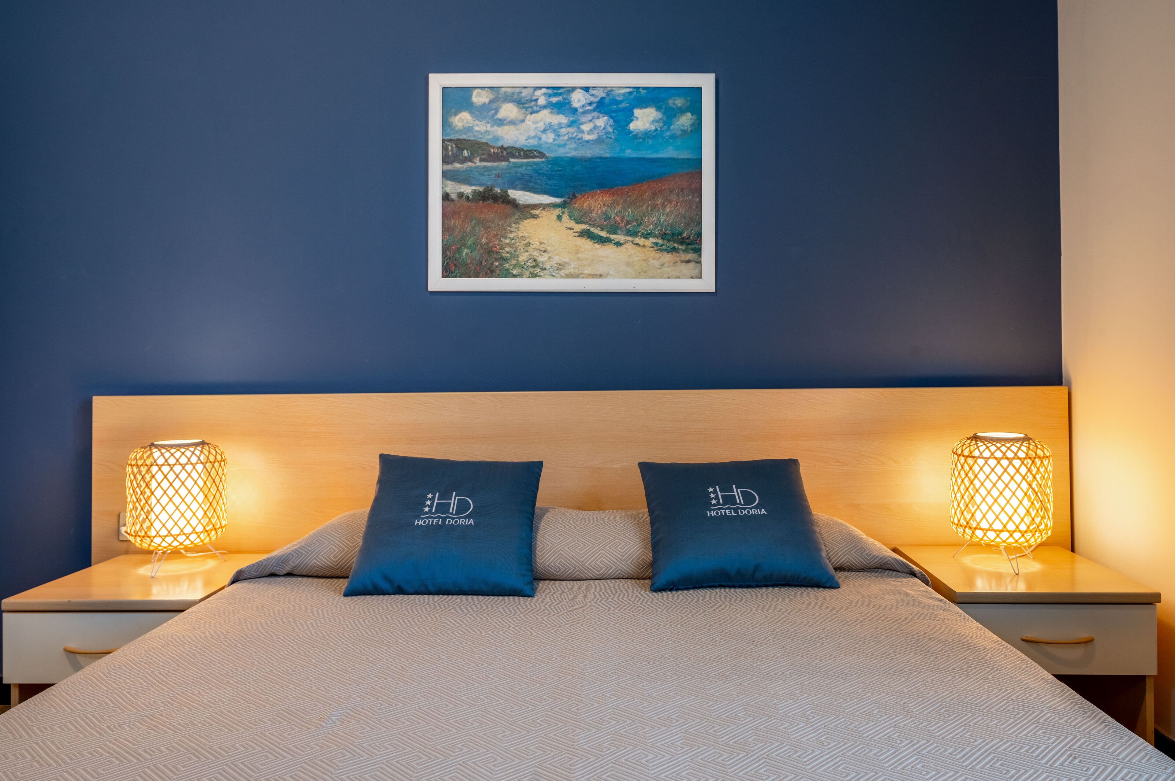 A hotel room with a bed, two nightstands with lamps, a painting on the wall, and pillows with the hotel logo.