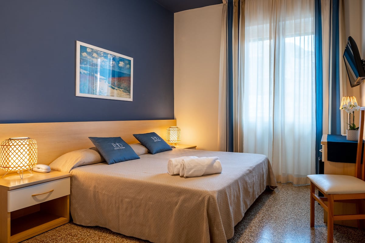 A hotel room with a bed, bedside tables, a lamp, a chair, a TV, and a window with curtains.