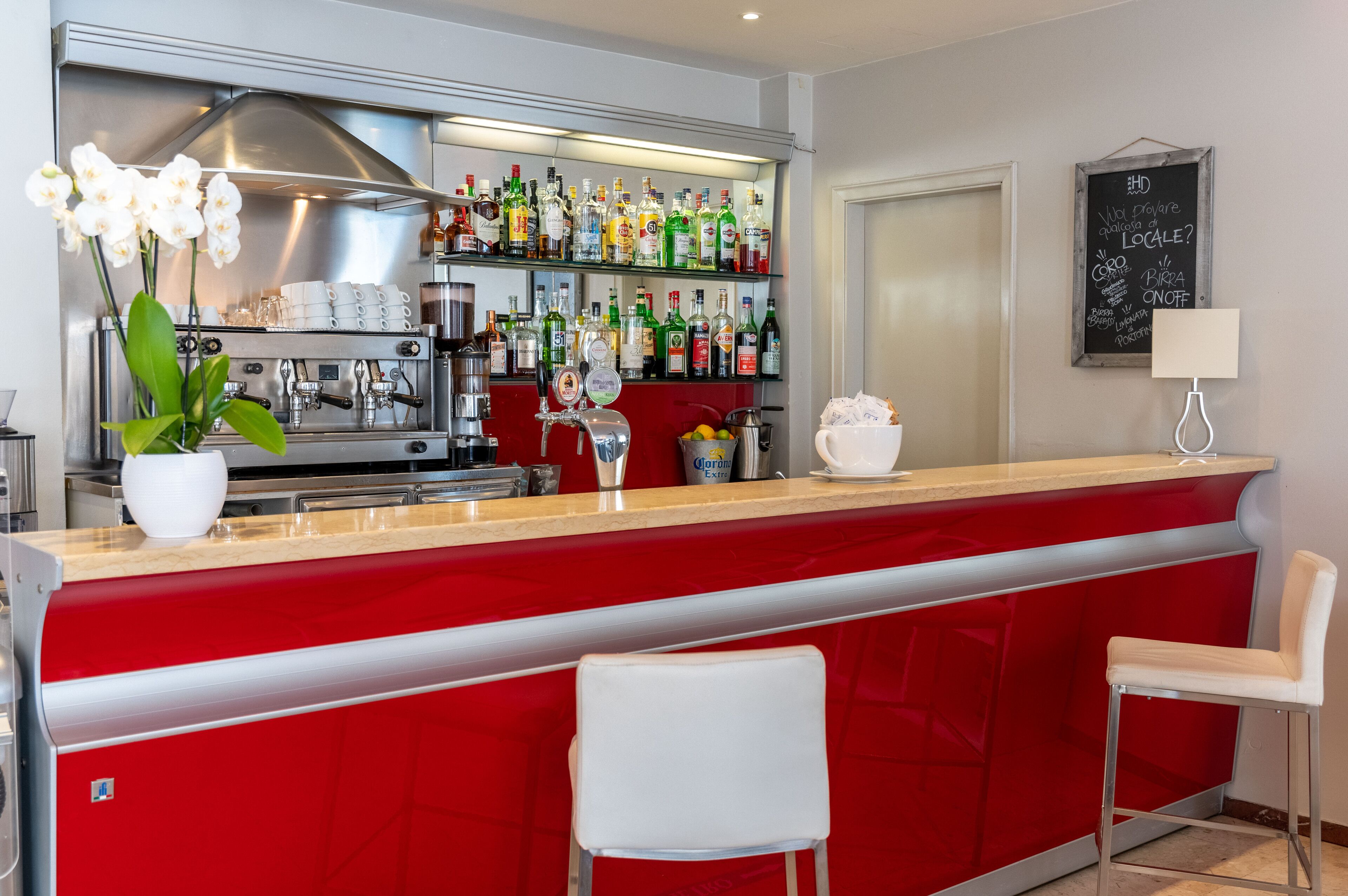 A bar area with a red counter, white chairs, a coffee machine, and shelves stocked with bottles.