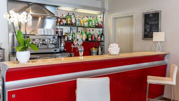 Zona de bar con mostrador rojo, sillas blancas, una máquina de café y estantes llenos de botellas.