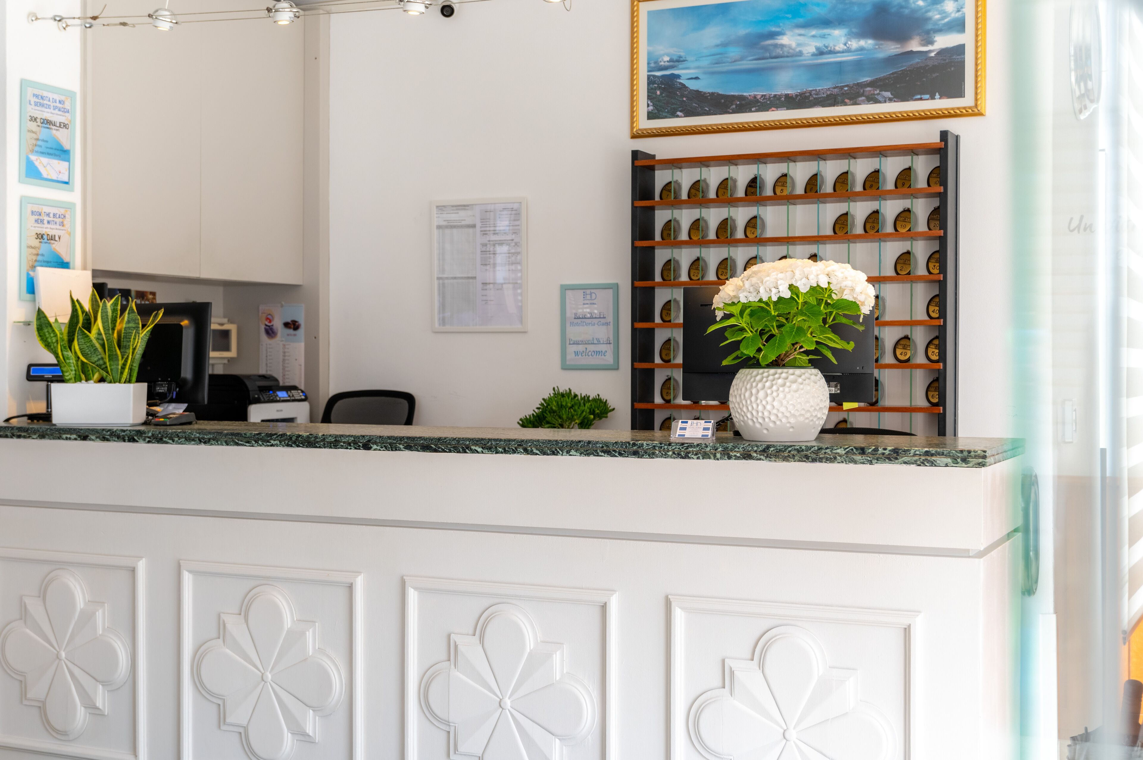 A reception area with a white counter, a potted plant, a framed picture on the wall, and a bulletin board with a welcome sign.
