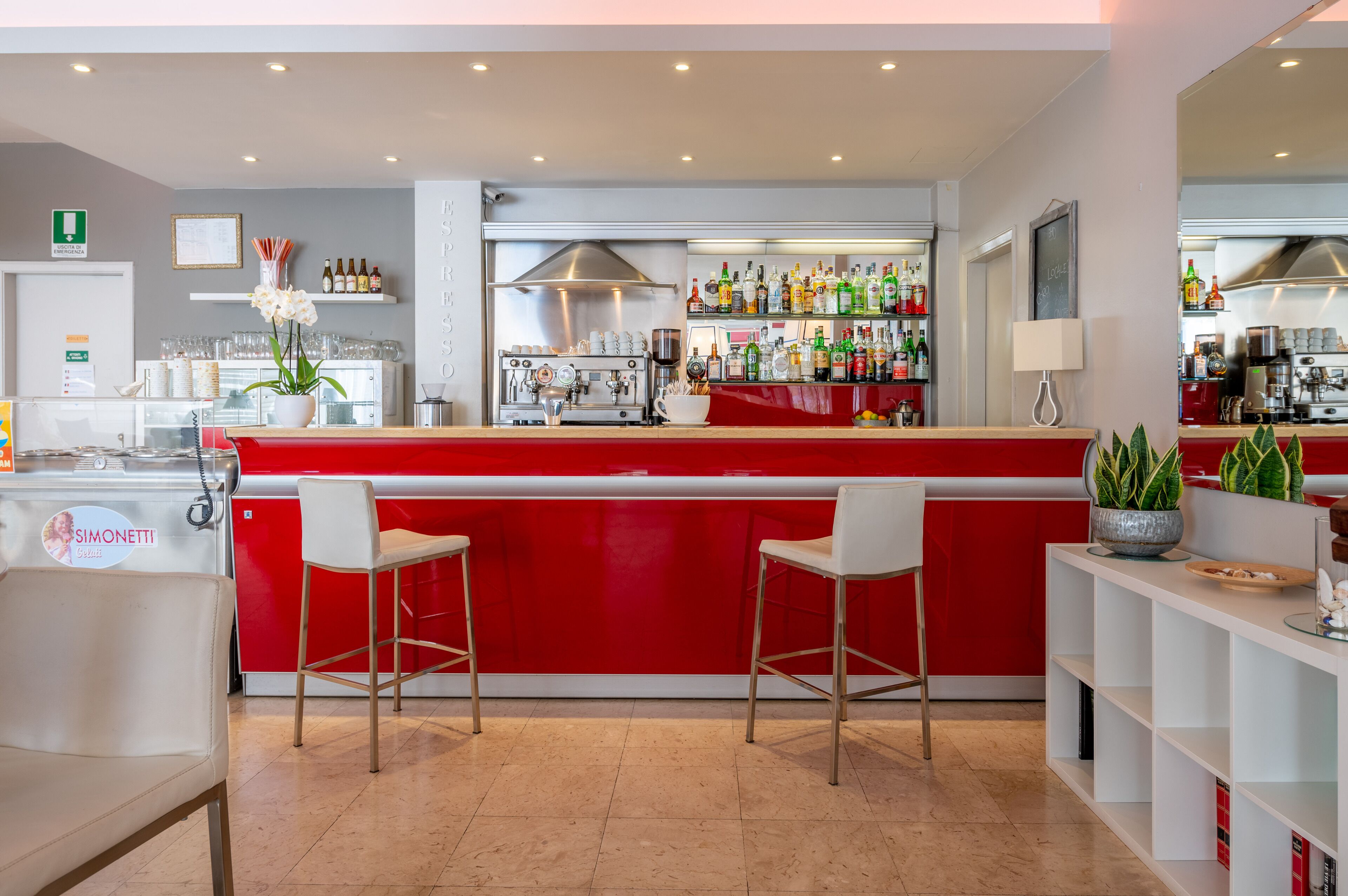 Un espace bar moderne avec un comptoir rouge, deux tabourets de bar blancs et une variété de bouteilles et de verres disposés sur des étagères.
