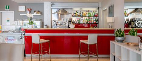 A modern bar area with red bar counter, two white bar stools, and a variety of bottles and glasses on shelves.
