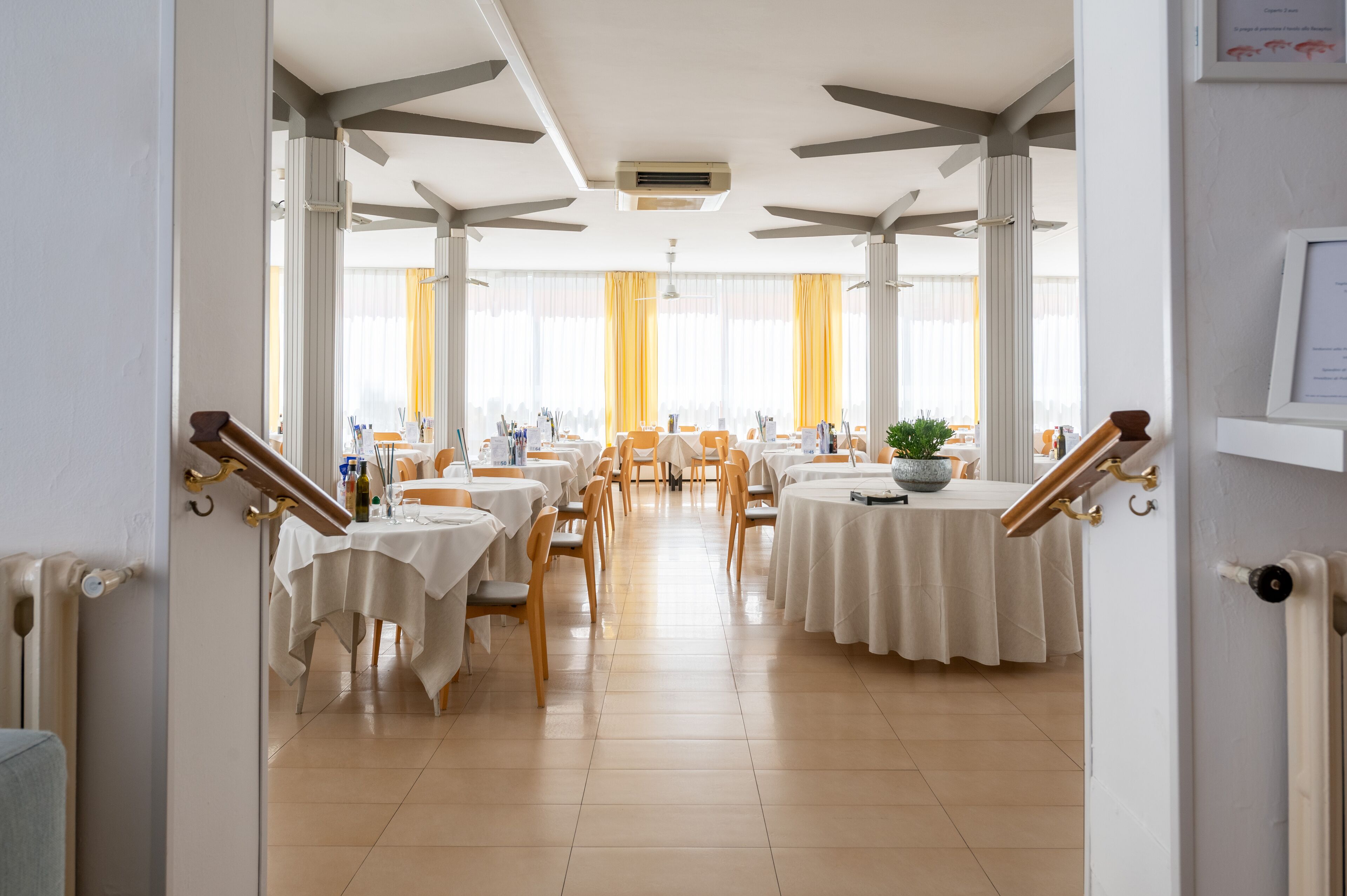 A dining area with tables set for service, a staircase with a handrail, and a view of the dining area through a glass door.