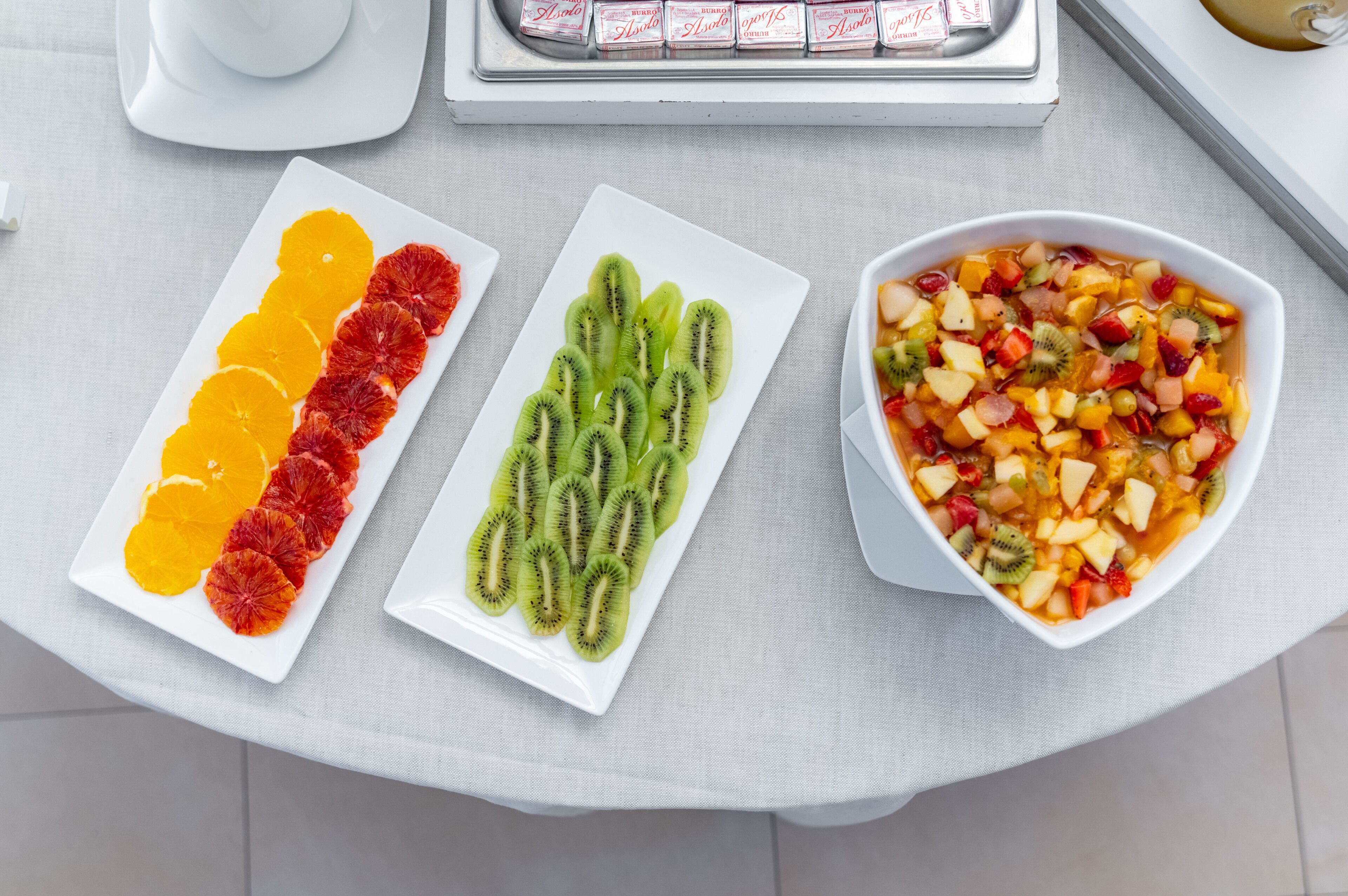 Ein Tisch mit drei weißen Schüsseln: eine mit Orangenscheiben, eine mit Kiwischnitten und eine mit Obstsalat.