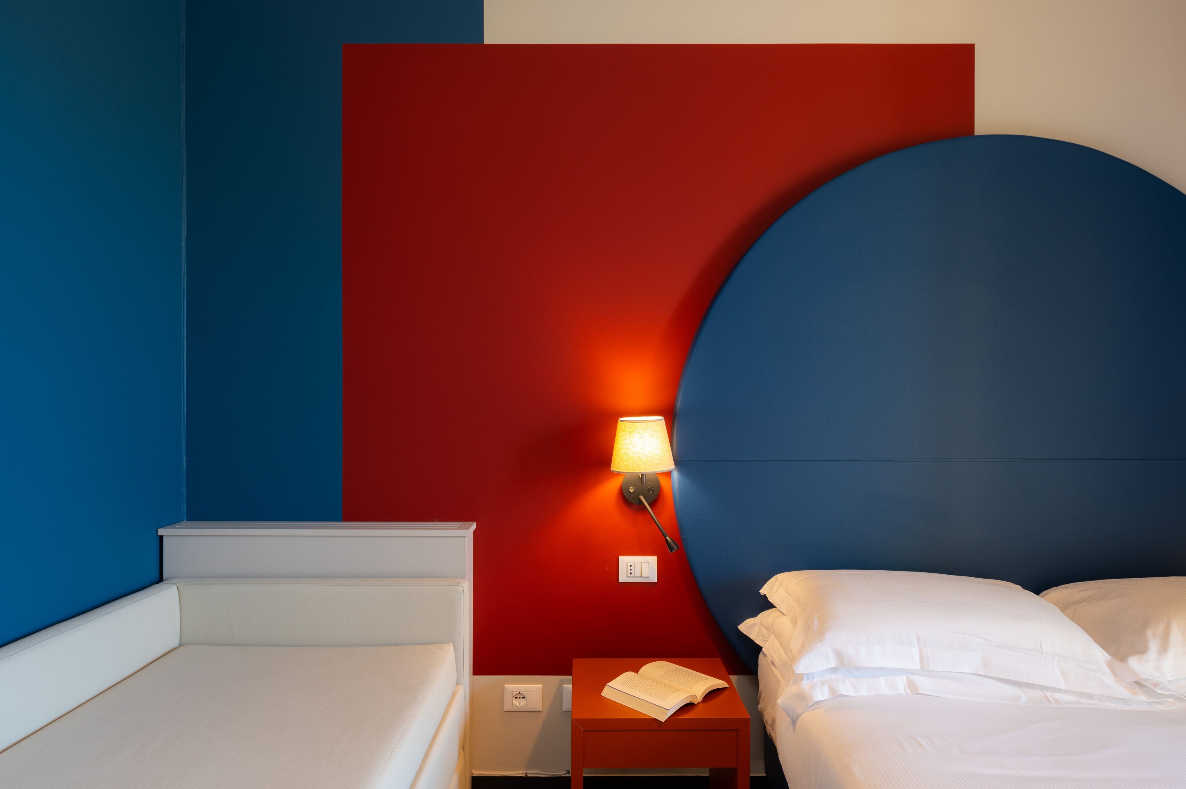 A hotel room with a bed, a nightstand, a lamp, and a red accent wall.