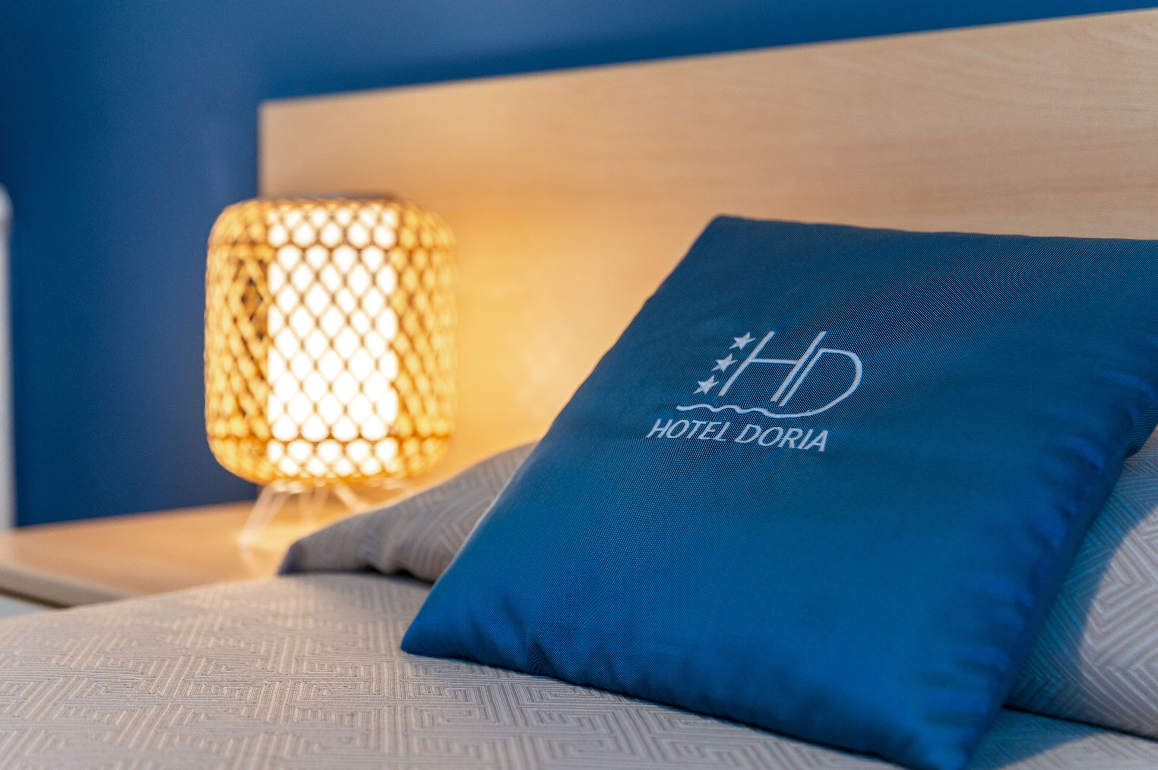 Een bed met een blauw kussen waarop het logo van Hotel Doria staat, een gestructureerde muur en een lamp met een honingraatmotief.