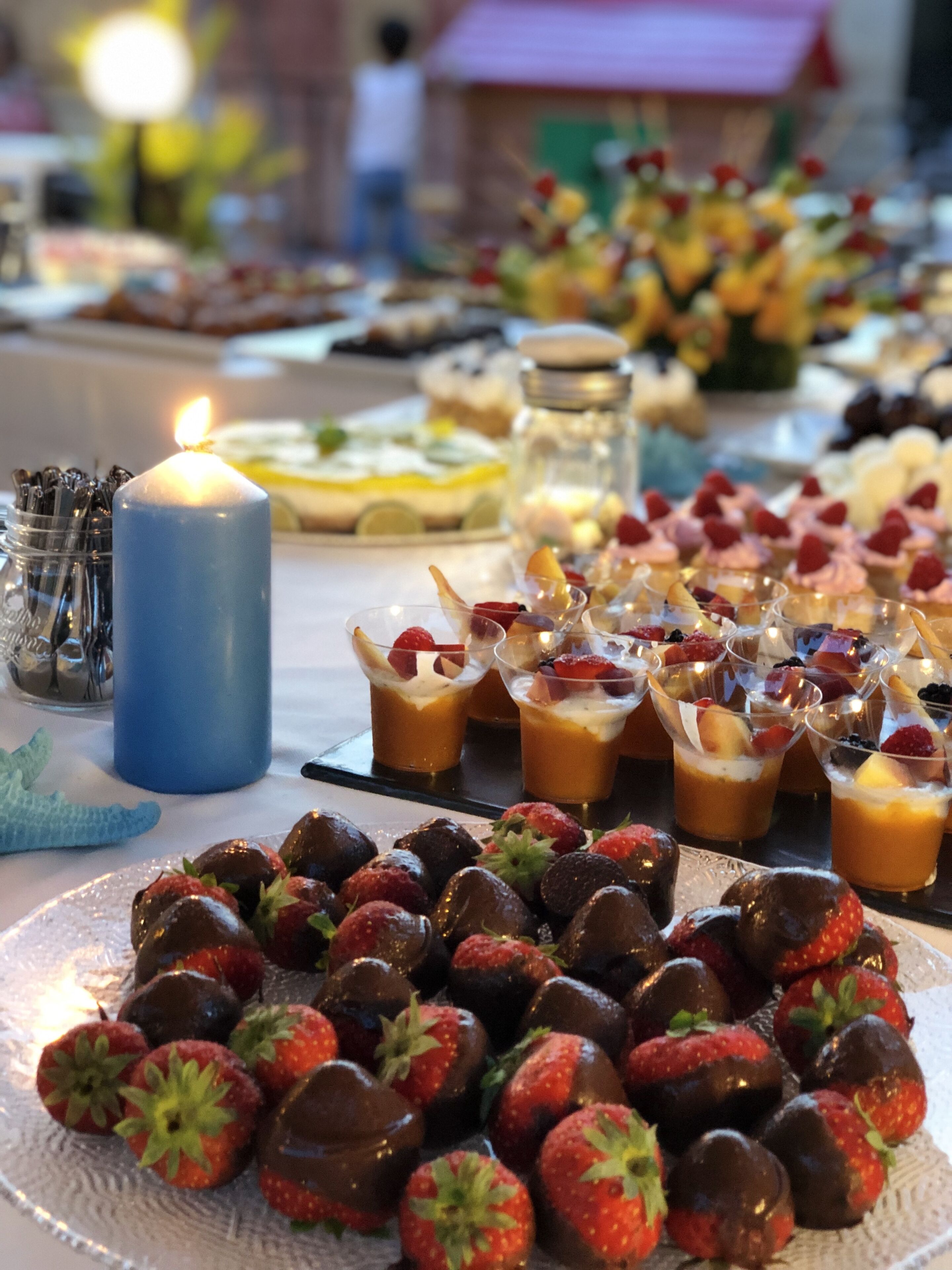 Una mesa de postres con fresas cubiertas de chocolate, vasos de fruta y una vela encendida.