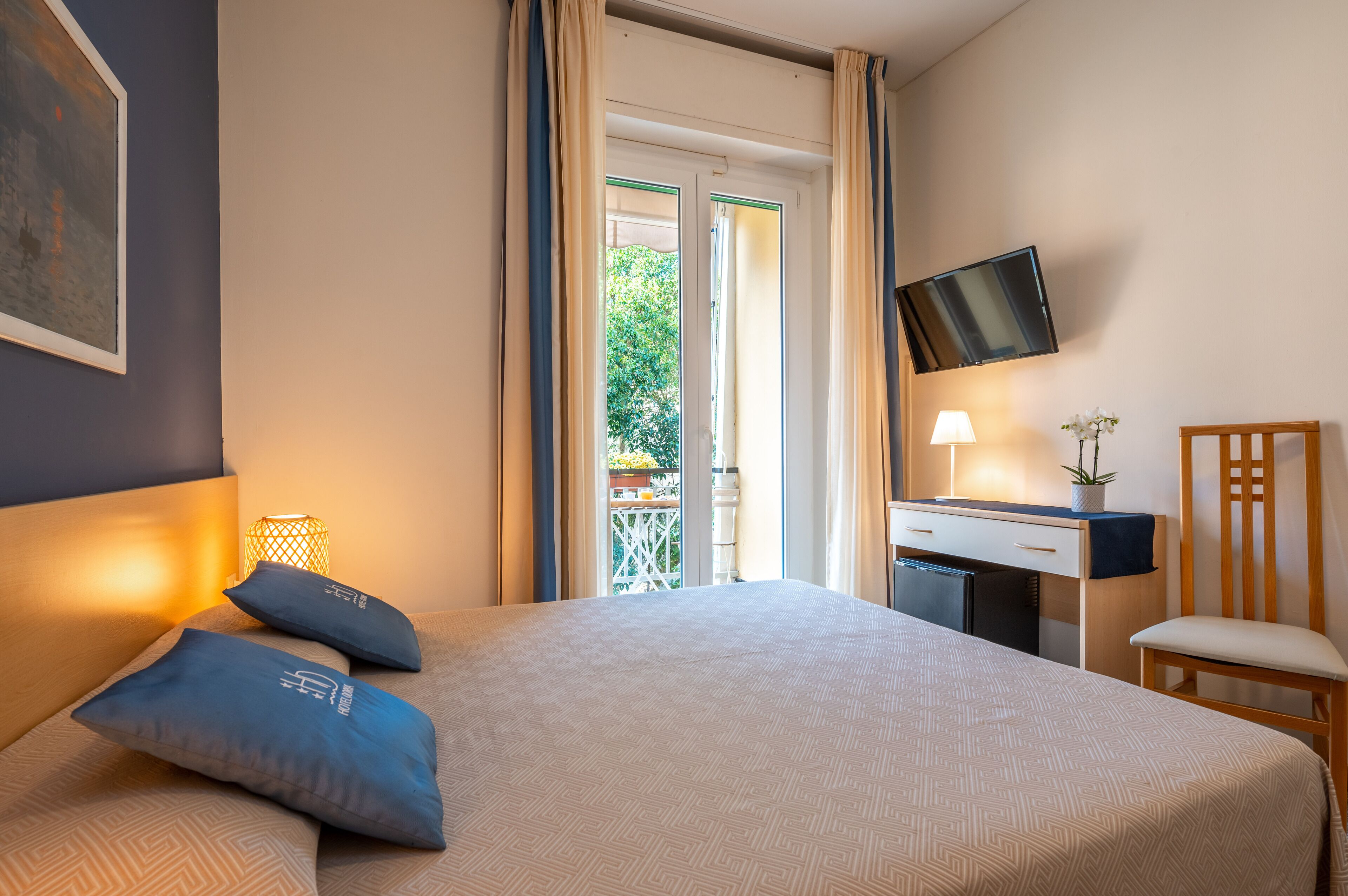 A hotel room with a bed, a desk, a chair, a TV, and a balcony with curtains.