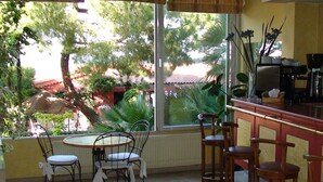 A dining area with a round table and chairs, a bar counter with stools, and a large window offering a view of trees and a building outside. - Hôtel Alexandra (Roquebrune-Cap-Martin)