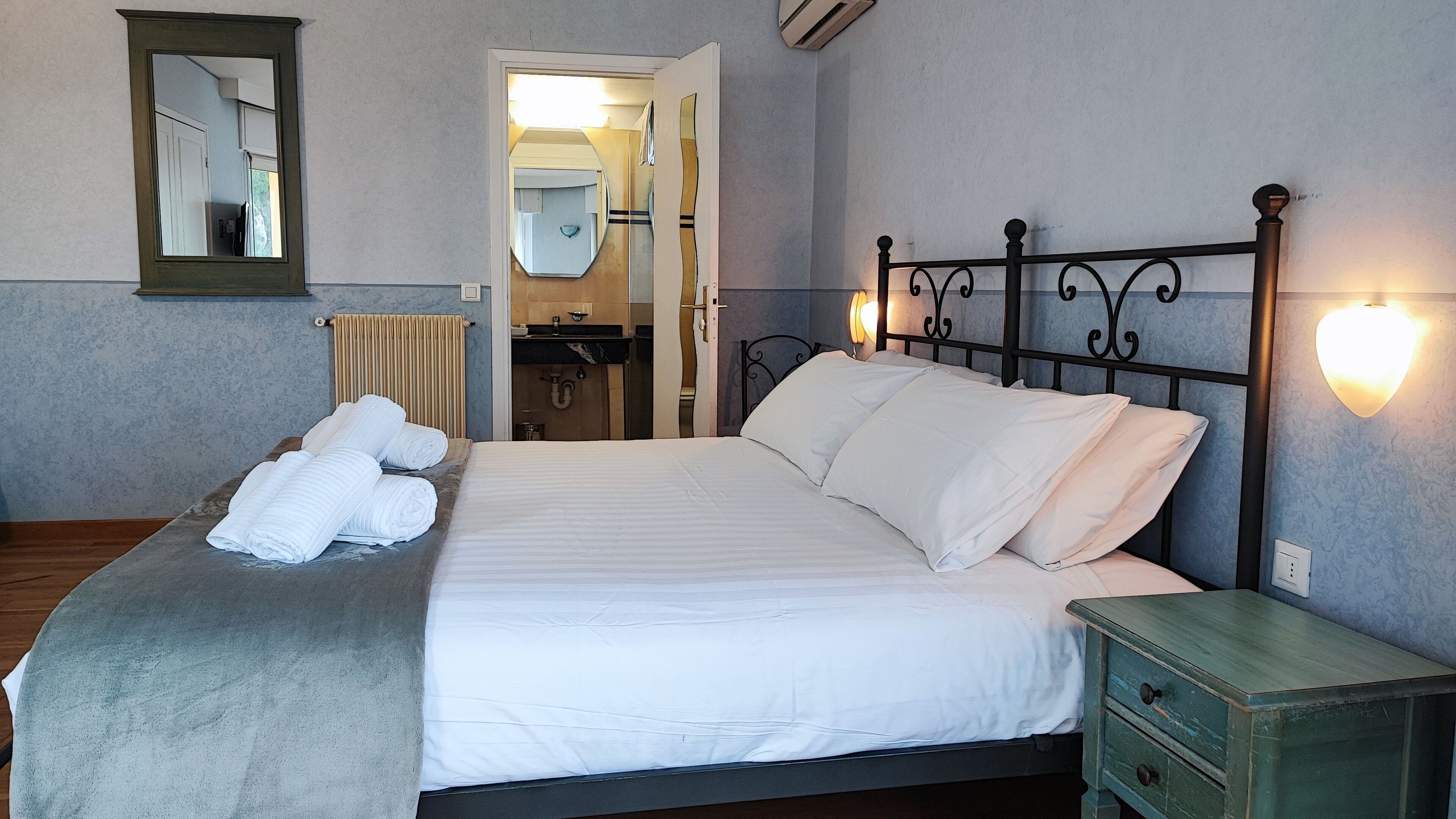 A neatly made bed with white linens and rolled towels, a bedside table with a green drawer, and a wall-mounted light fixture.