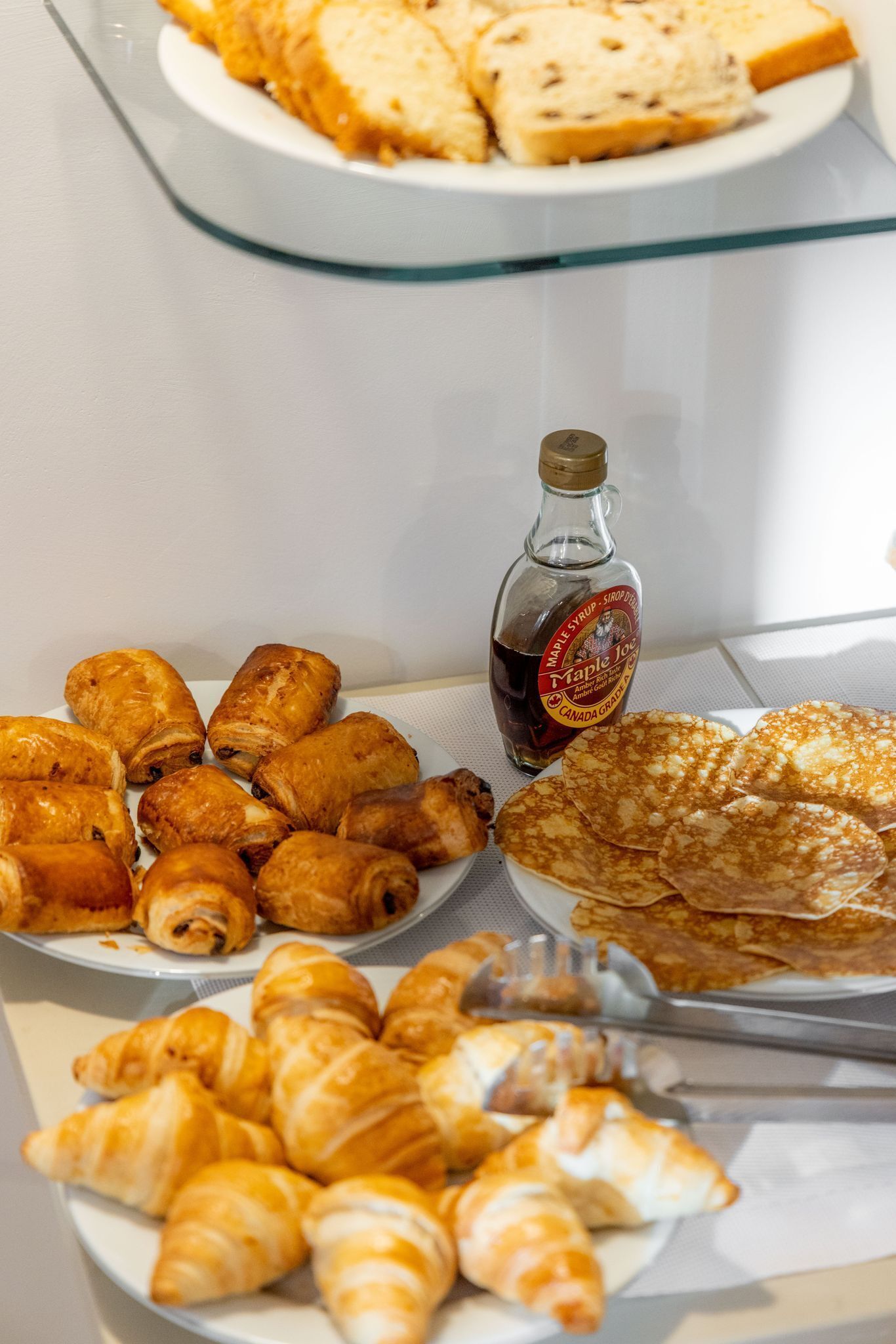 A buffet with various pastries, including croissants and rolls, and a bottle of maple syrup.