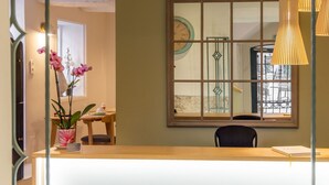 A reception area with a wooden desk, a chair, a vase with flowers, a clock, and a window with a view. - Hotel Les Armoiries (Valbonne)