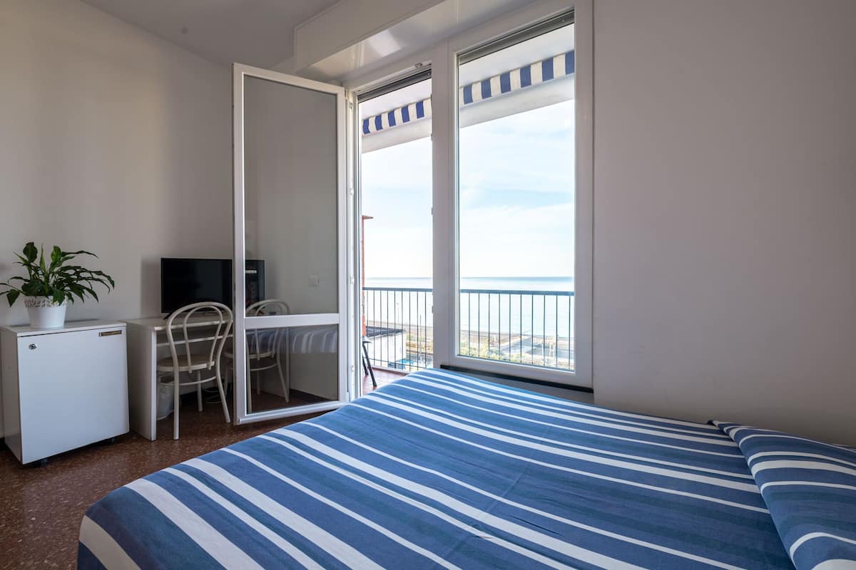 A bedroom with a bed, a desk, a chair, a small fridge, and a balcony with a view of the sea.