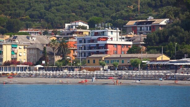 Un hotel frente al mar con varios pisos, una playa en primer plano y un fondo de árboles y edificios.