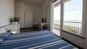 Un dormitorio con cama, escritorio, silla, armario y balcón con vista al mar.