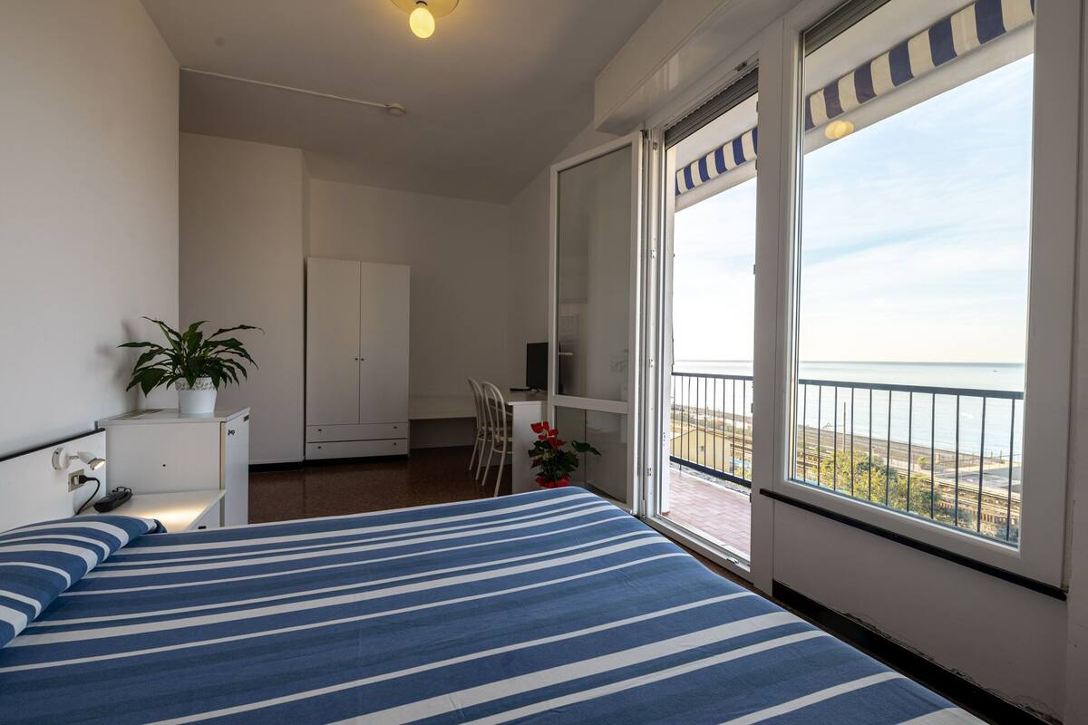 A bedroom with a bed, a desk, a chair, a wardrobe, and a balcony with a view of the sea.
