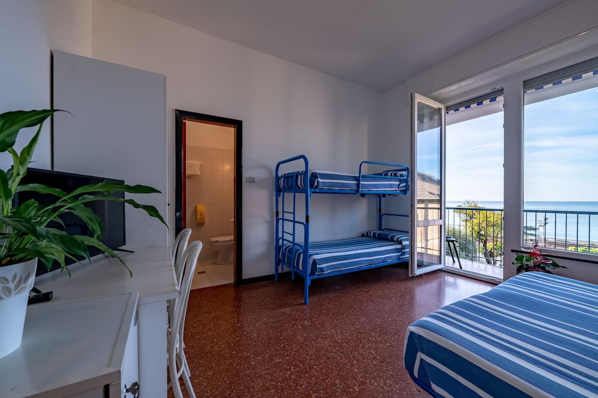 A hotel room with a bunk bed, a desk with a chair, a potted plant, and a large window offering a view of the sea.