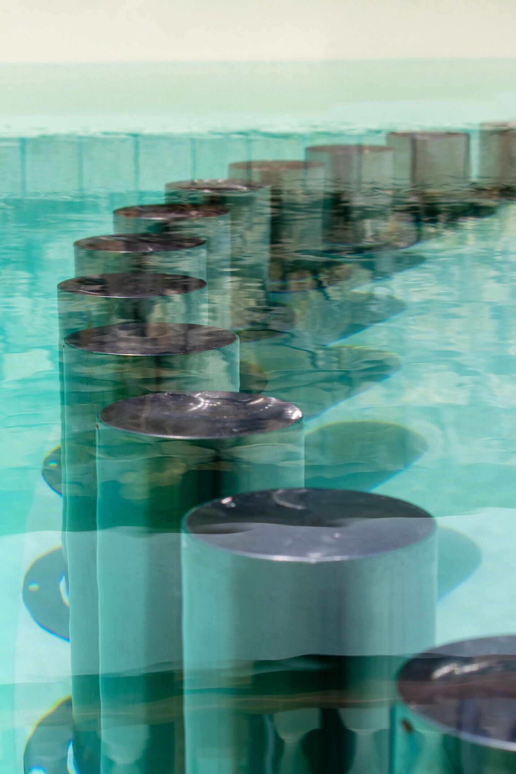 Zylindrische Objekte spiegeln sich in einer reflektierenden Oberfläche, möglicherweise einem Pool oder einer Wasserspiele-Anlage.