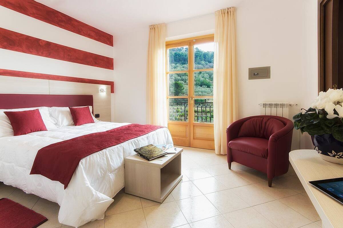 Habitación de hotel con una cama grande, un sillón rojo, una mesita con un libro y una ventana que ofrece vistas a zonas verdes.