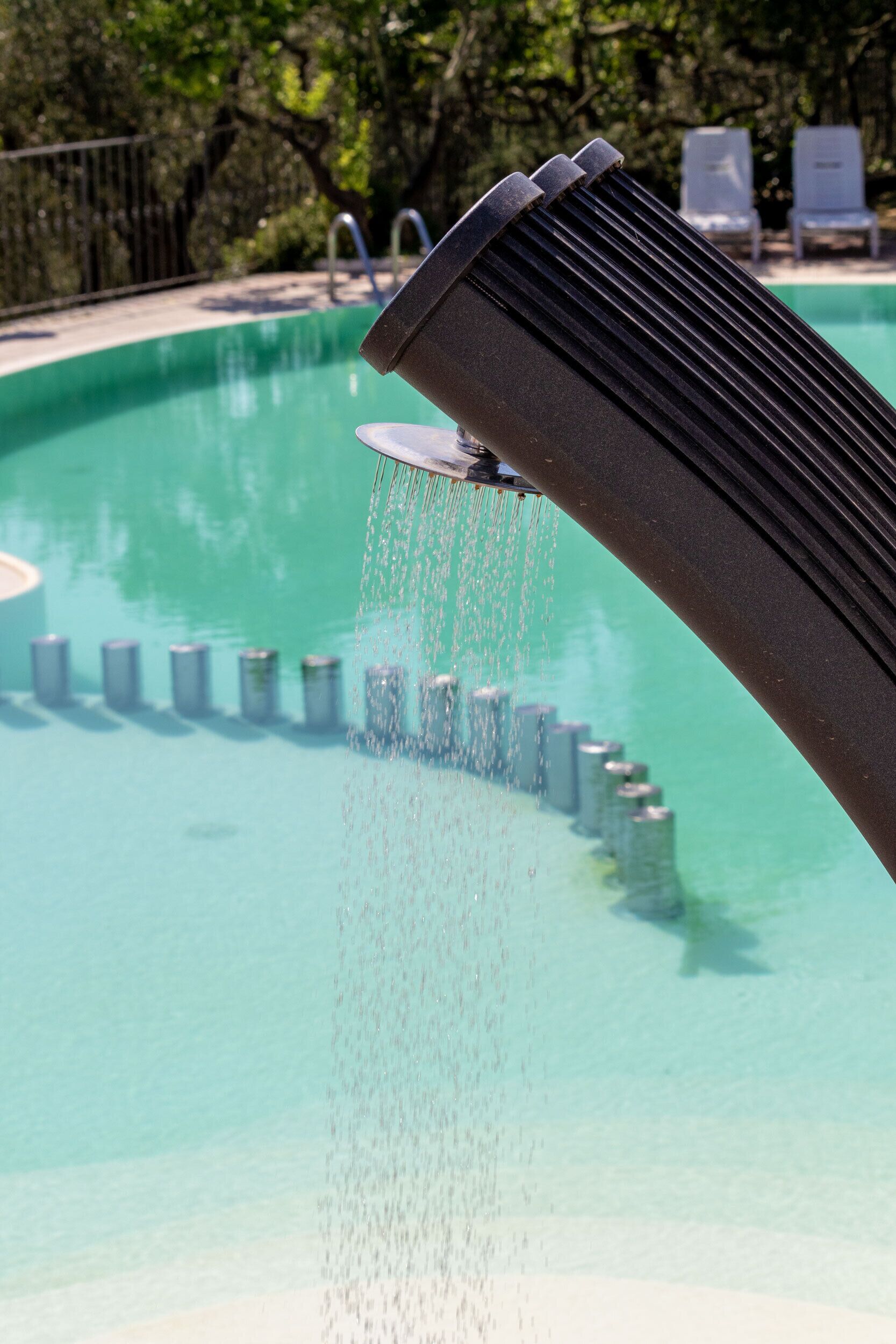A water feature with a series of cylindrical objects arranged in a semi-circle, a pool, and a metal railing in the background.