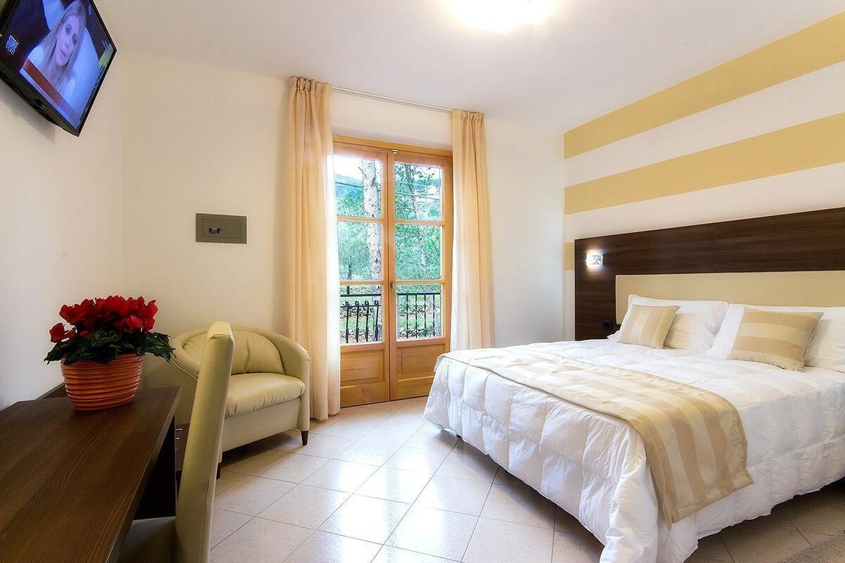 Habitación de hotel con cama, una silla, televisor, ventana con cortinas y una planta en maceta.