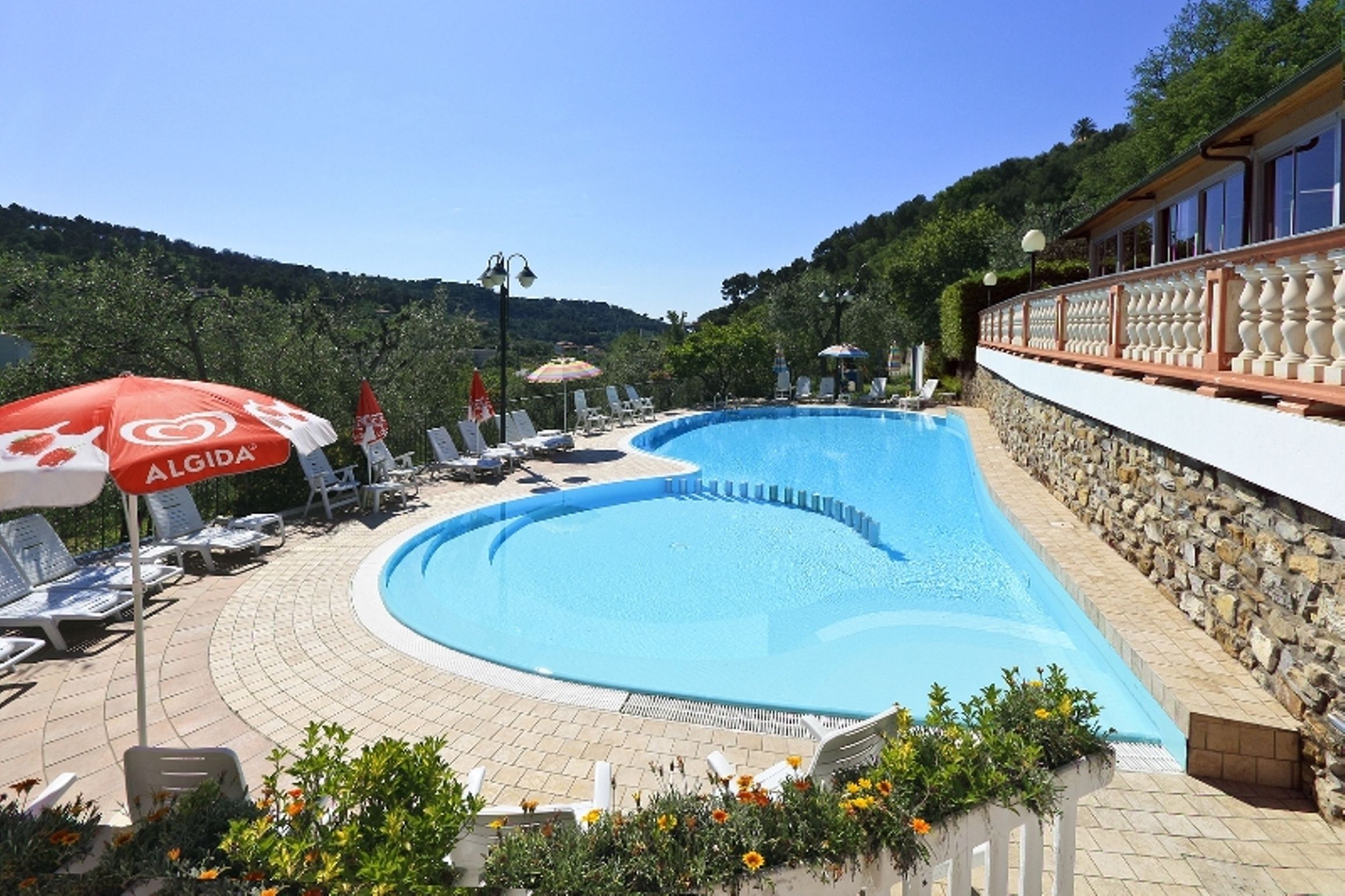 A swimming pool with lounge chairs and umbrellas, surrounded by a stone wall and greenery.