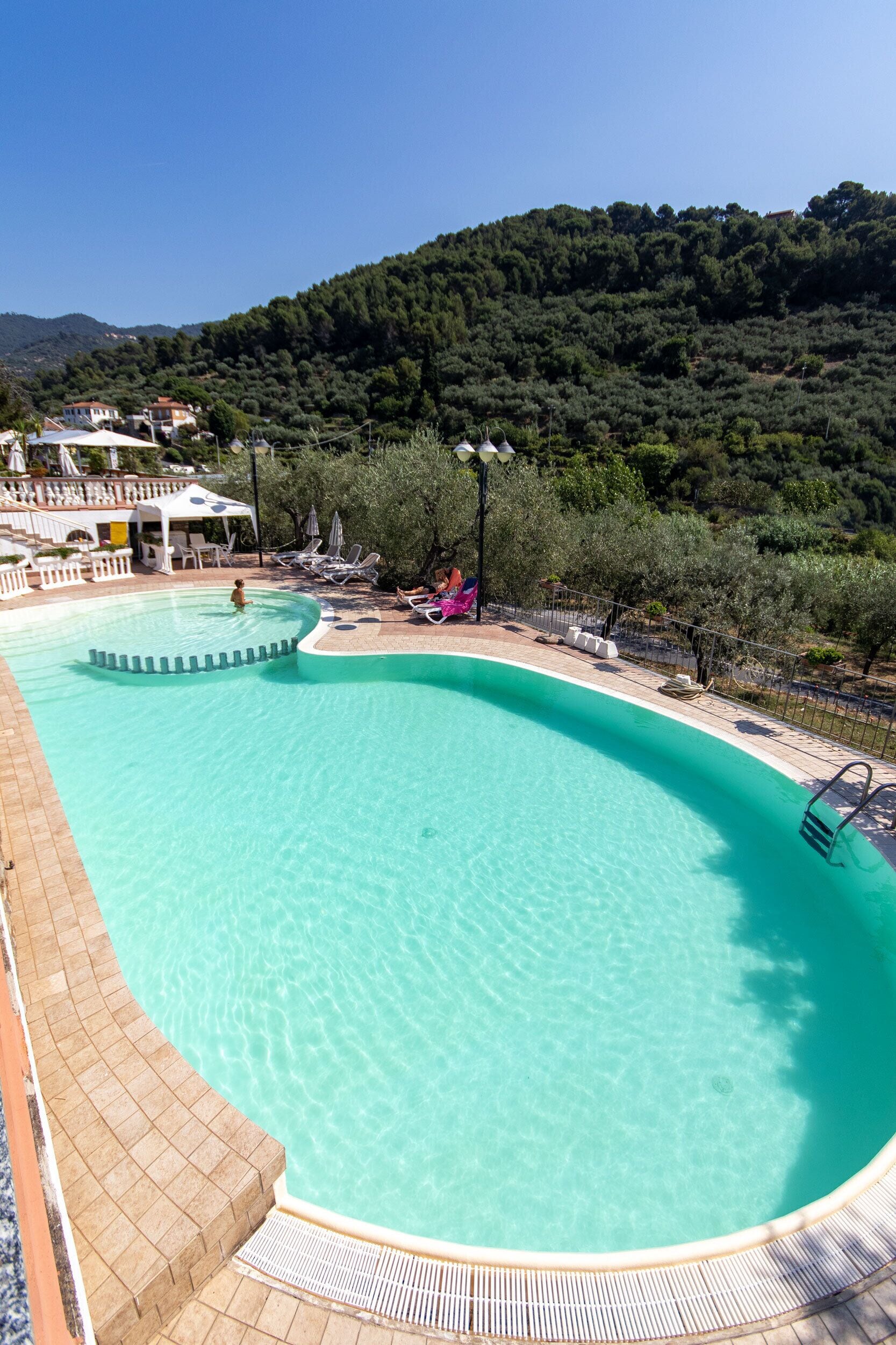 A large, curved swimming pool with clear water, surrounded by lounge chairs and umbrellas, set against a backdrop of lush greenery and hills.