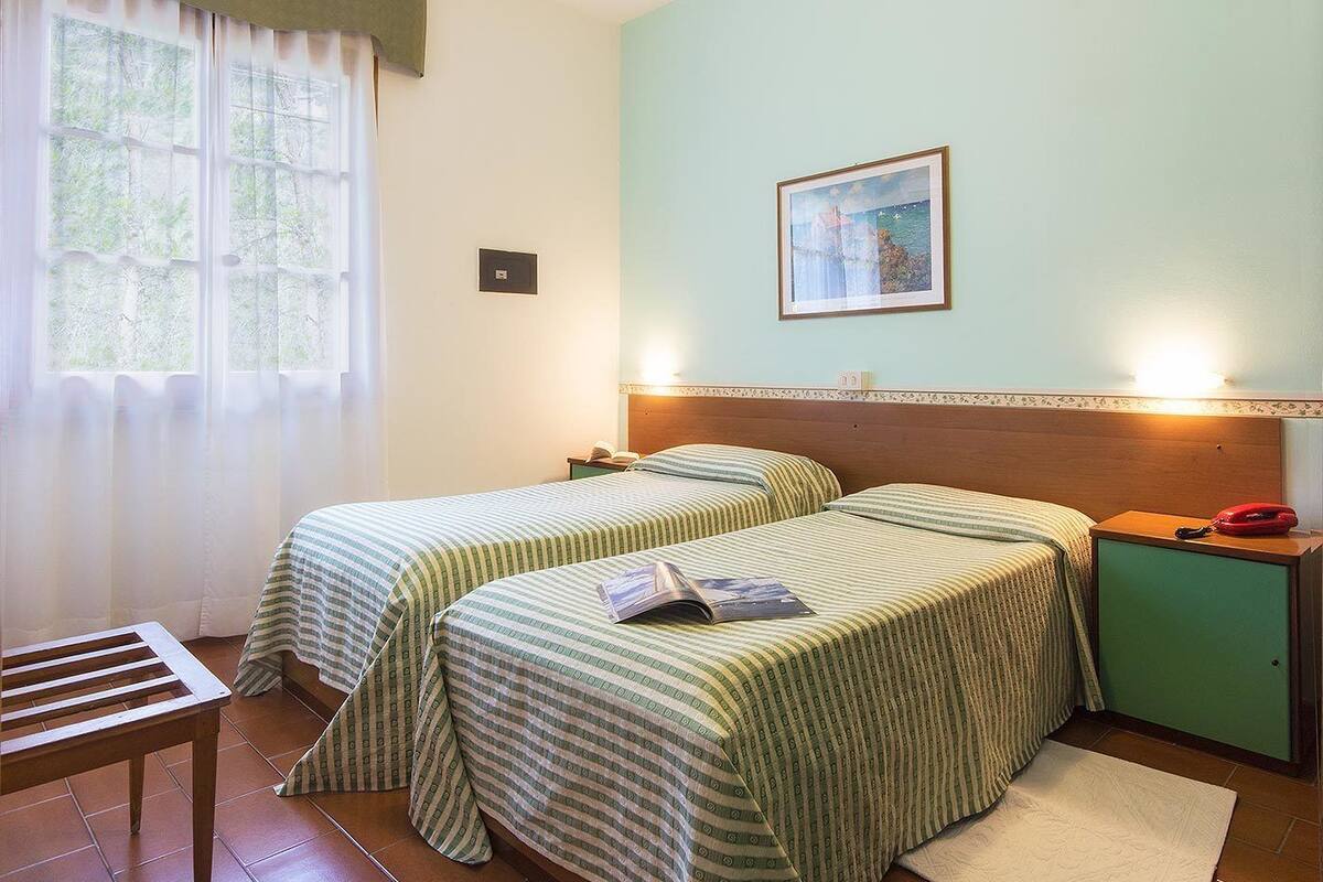 Habitación con dos camas, cabecero de madera, mesita de noche con lámpara, un armario pequeño y una ventana con cortinas.