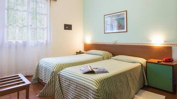 A room with two beds, a wooden headboard, a bedside table with a lamp, a small cabinet, and a window with curtains.