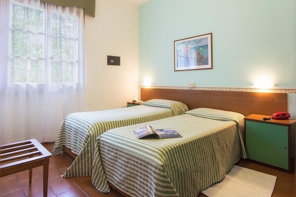 Une chambre avec deux lits, une tête de lit en bois, une table de chevet avec une lampe, une petite armoire et une fenêtre avec des rideaux.